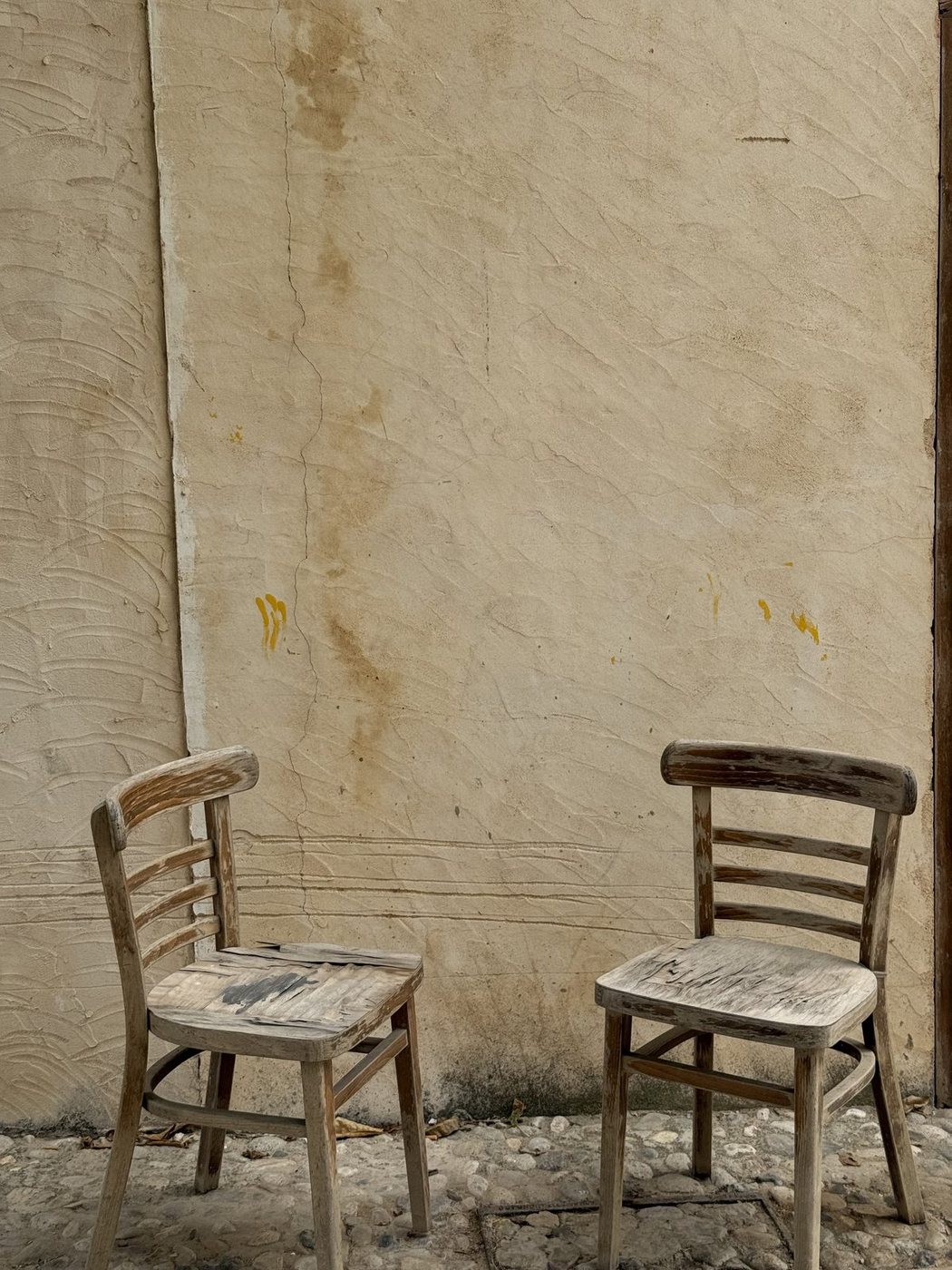 Two weathered wooden chairs against a sunlit plaster wall